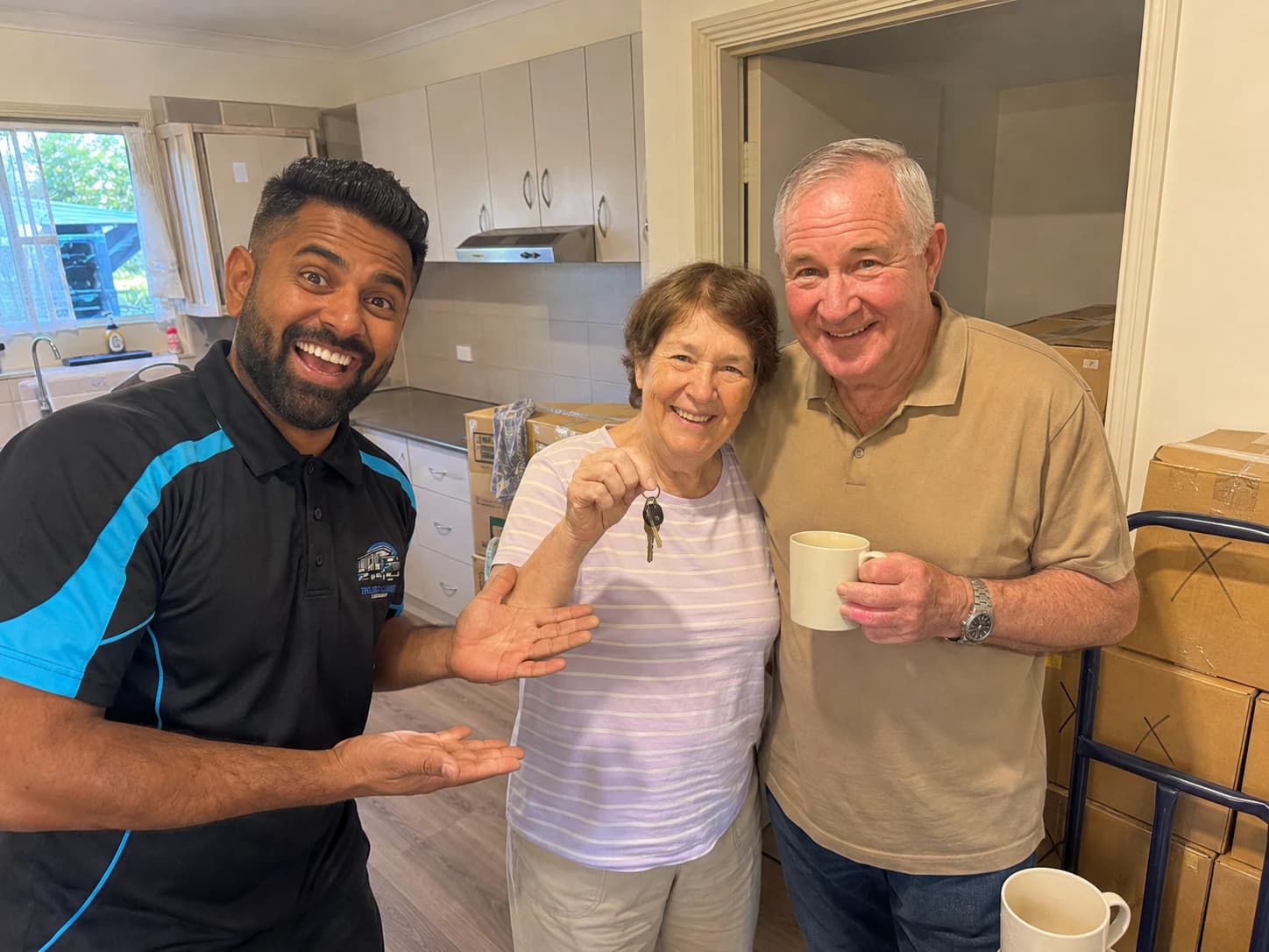 A Load Lounge crew member in the new kitchen with an older couple, the keys held up between them and packed boxes still stacked against the wall