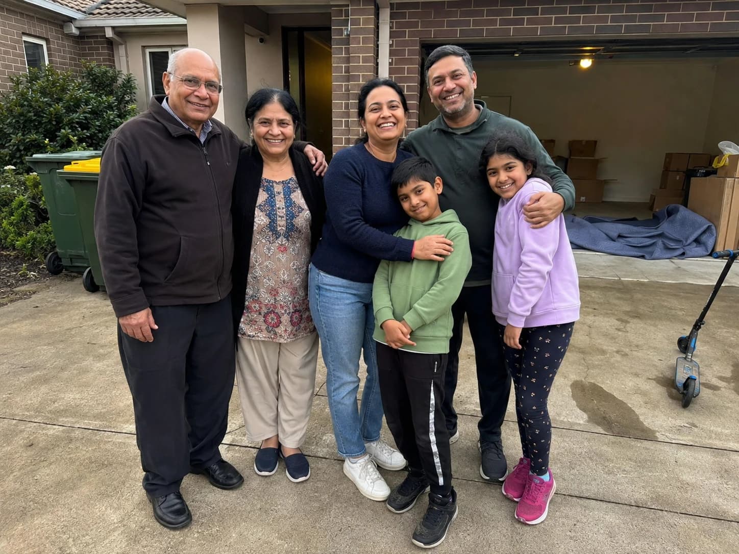 A family together on the driveway of their new home, the garage behind them stacked with boxes and moving blankets